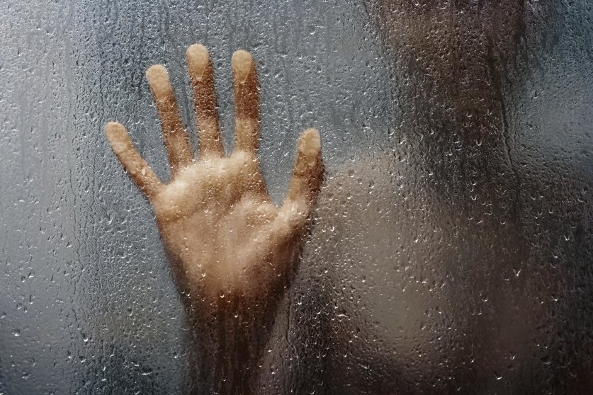 human hand and unrecognizable silhouette behind wet glass in shower 