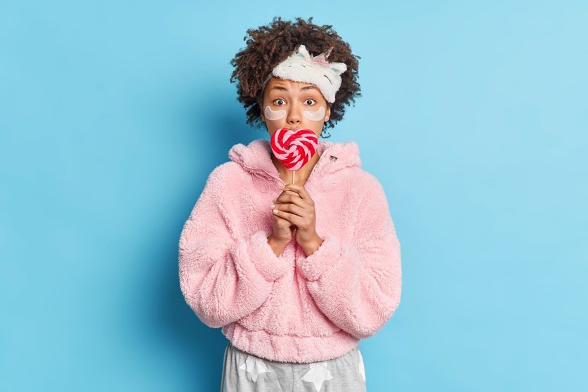 Surprised dark skinned woman has curly hair covers mouth with delicious candy wears sleepmask and pajama applies collagen patches under eyes to reduce wrinkles isolated over blue background.
