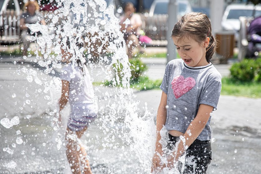 Funny little girl in a fountain, among the splashes of water on a hot summer day.